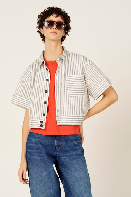 Blauw hemd met korte mouwen van Le Fabuleux Marcel de Bruxelles, met witte strepen, gedragen over een oranje T-shirt en blauwe jeans.
