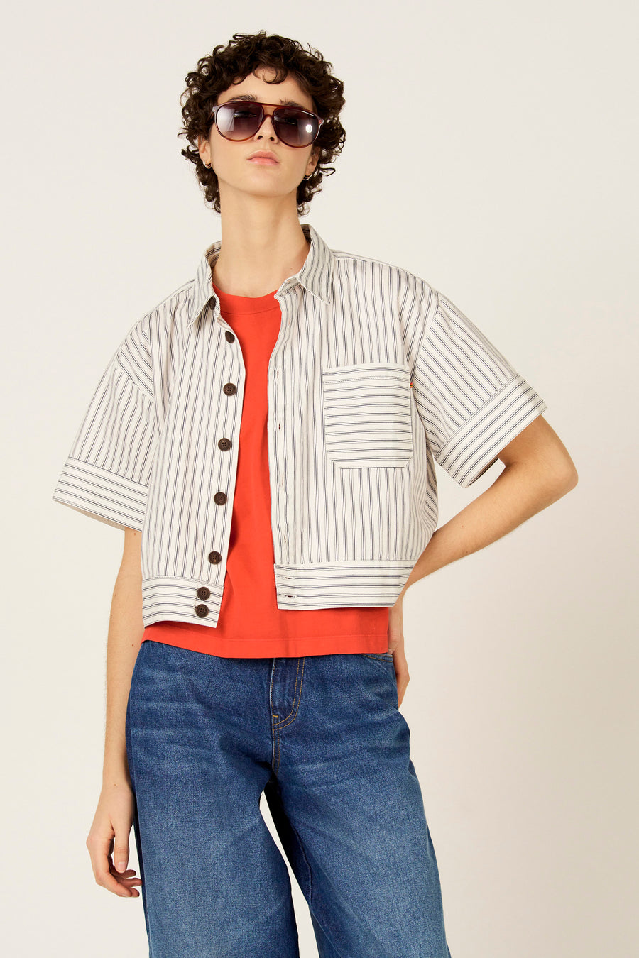 Blauw hemd met korte mouwen van Le Fabuleux Marcel de Bruxelles, met witte strepen, gedragen over een oranje T-shirt en blauwe jeans.
