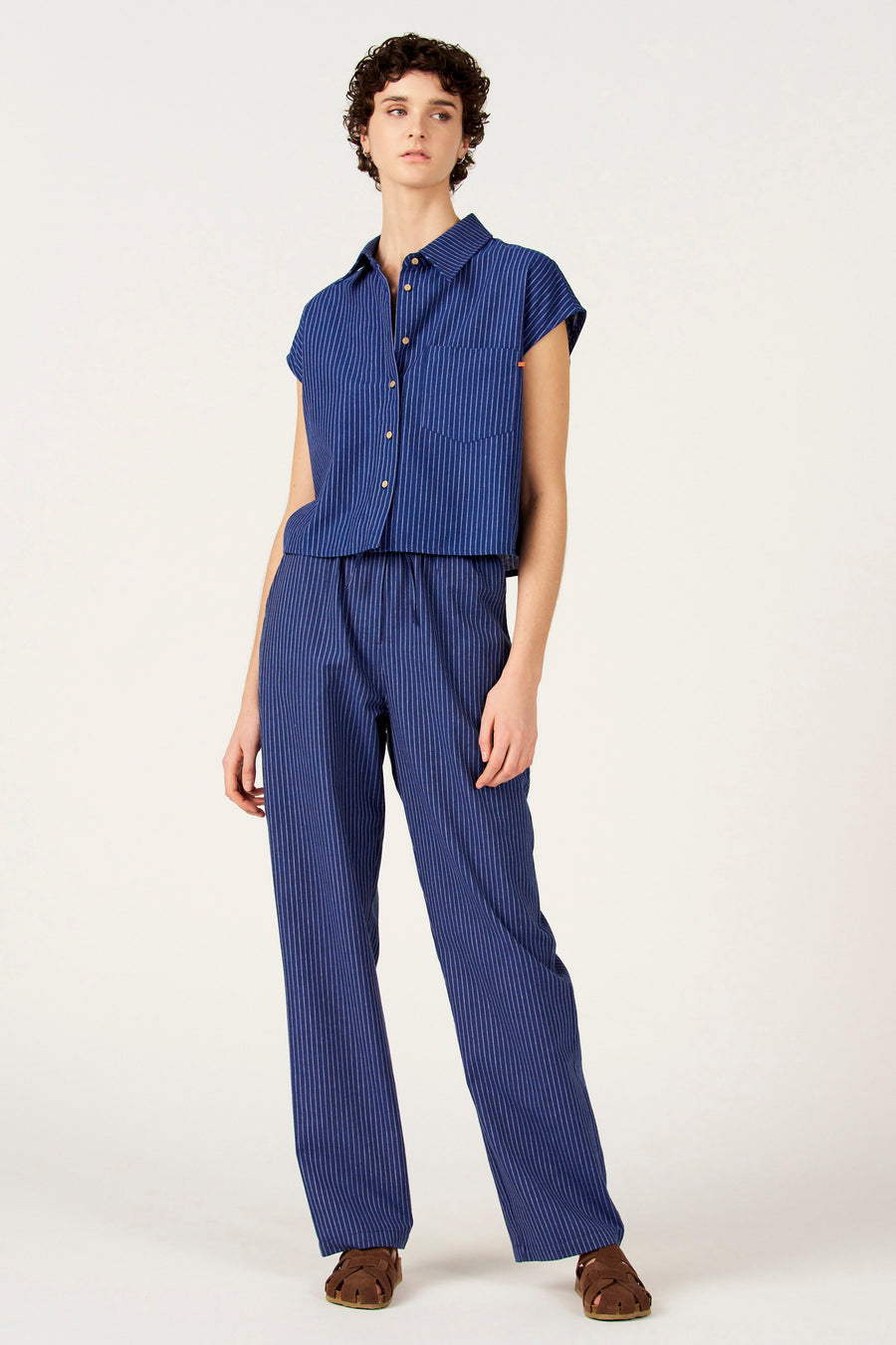Chemise bleue à rayures à manches courtes de Le Fabuleux Marcel de Bruxelles, avec boutons en bois et une poche poitrine, combinée avec un pantalon assorti à rayures et des sandales marron.