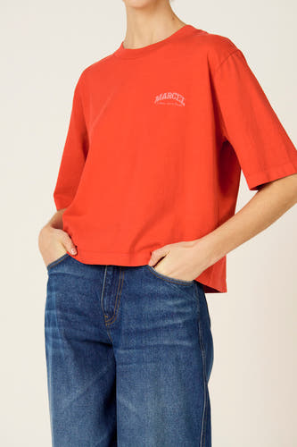 T-shirt rouge à manches courtes de Le Fabuleux Marcel de Bruxelles, avec un logo sur la poitrine et associé à un jean bleu.