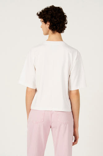 T-shirt blanc à manches courtes de Le Fabuleux Marcel de Bruxelles, avec un pantalon rose et des cheveux courts et bouclés.