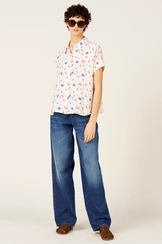 Chemise multicolore à manches courtes de Le Fabuleux Marcel de Bruxelles, avec un motif joyeux sur le thème de la plage. Portée avec un jean à jambes larges et des sandales marron.
