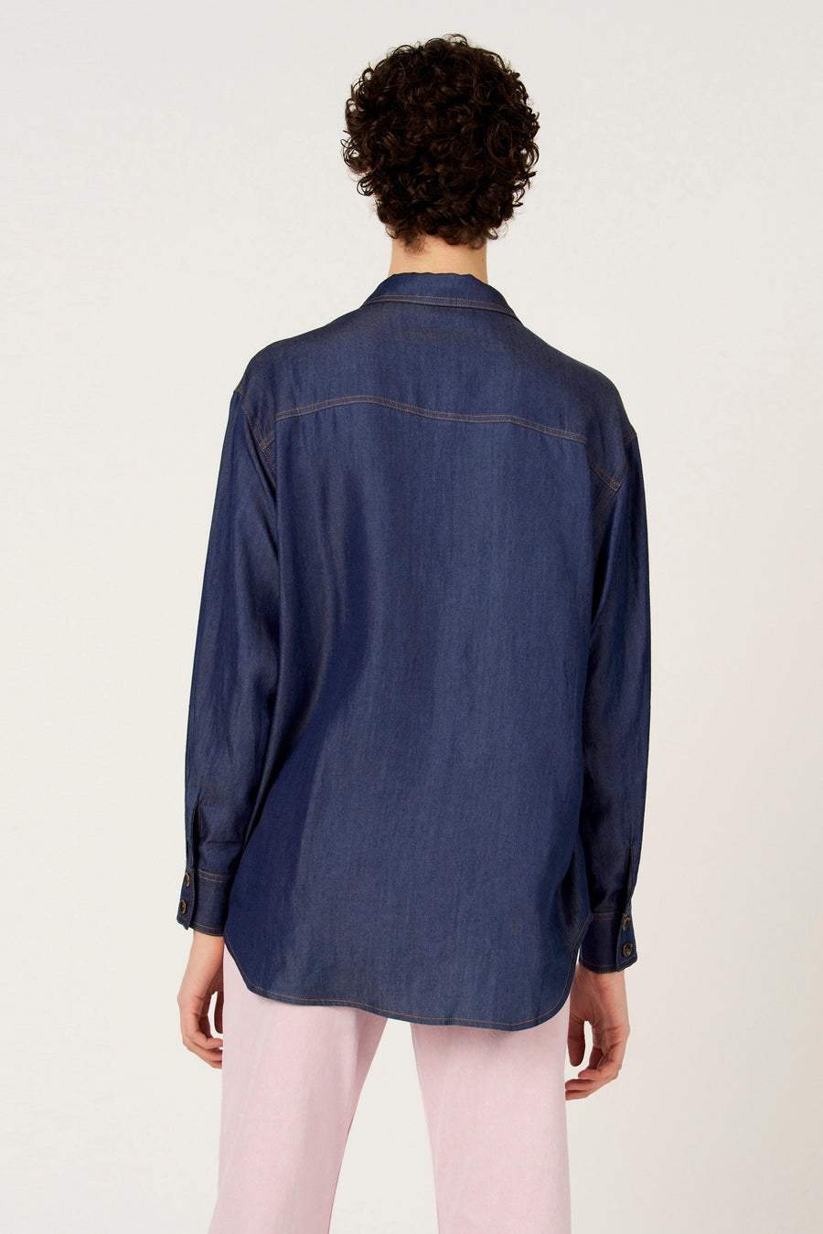 Chemise en jean bleu foncé à manches longues de Le Fabuleux Marcel de Bruxelles, vue de dos, avec surpiqûres marron et pantalon rose.