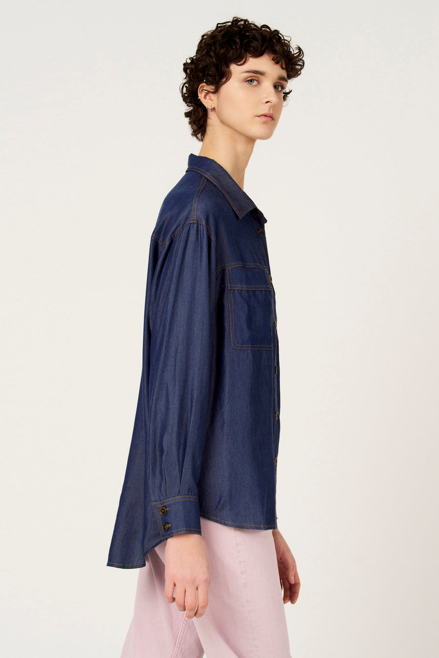 Chemise en jean bleu foncé à manches longues de Le Fabuleux Marcel de Bruxelles, avec poche poitrine et fermeture boutonnée, portée avec un pantalon rose.