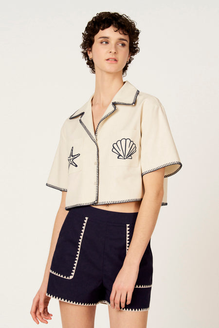 Chemise écru à manches courtes de Le Fabuleux Marcel de Bruxelles, finie avec des surpiqûres contrastées, portée avec un short bleu foncé.