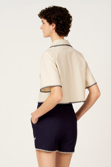 Ecru hemd met korte mouwen van Le Fabuleux Marcel de Bruxelles, afgewerkt met contrasterende stiksels, gedragen met donkerblauwe shorts.
