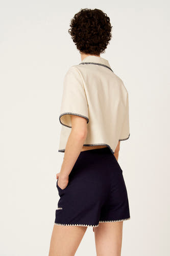 Chemise écru à manches courtes de Le Fabuleux Marcel de Bruxelles, avec détails zigzag contrastants, portée avec un short bleu foncé.