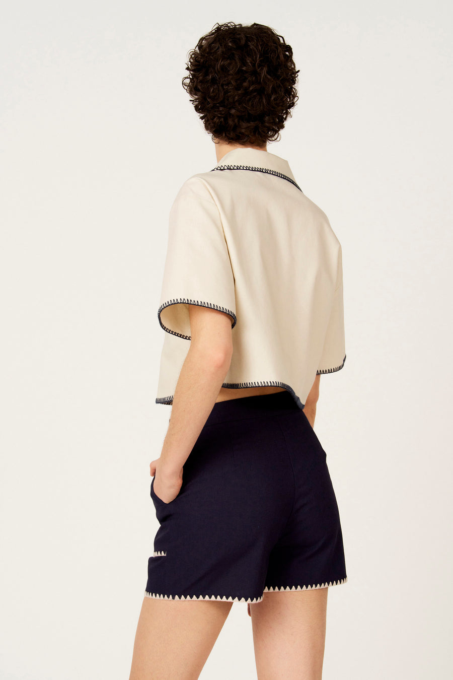 Chemise écru à manches courtes de Le Fabuleux Marcel de Bruxelles, avec détails zigzag contrastants, portée avec un short bleu foncé.