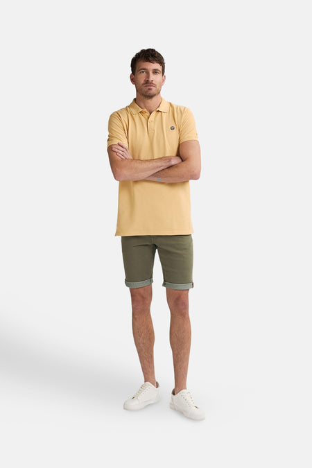 Gele polo met korte mouwen van Petrol Industries, met gekruiste armen en groene shorts gecombineerd met witte schoenen.