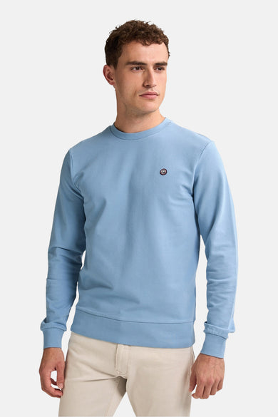 Man in lichtblauw sweatshirt met logo en beige broek.

