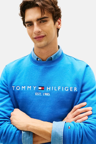 Pull bleu à col rond de TOMMY HILFIGER, avec imprimé et manches longues, porté sur une chemise en jean.