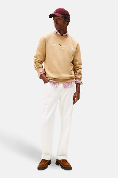 Gilet beige de TOMMY HILFIGER, avec un logo blanc et porté sur une chemise rayée, avec un chapeau bordeaux et un pantalon blanc.