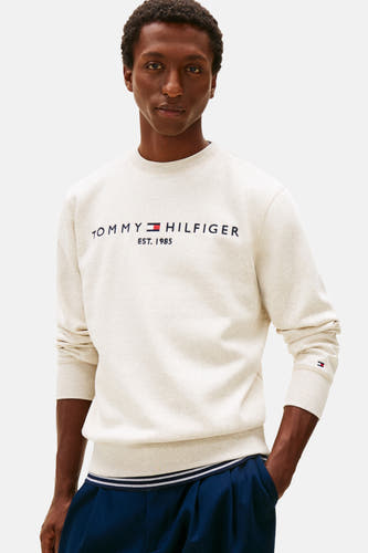 Gilet beige de TOMMY HILFIGER avec logo de marque sur la poitrine, associé à un pantalon bleu et des baskets blanches.
