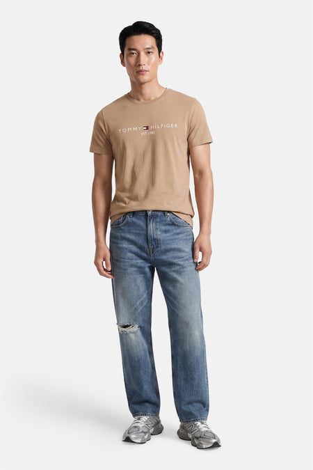 Beige korte mouwen T-shirt van TOMMY HILFIGER met wit logo op borst, gedragen met blauwe jeans en grijze sneakers, front view.
