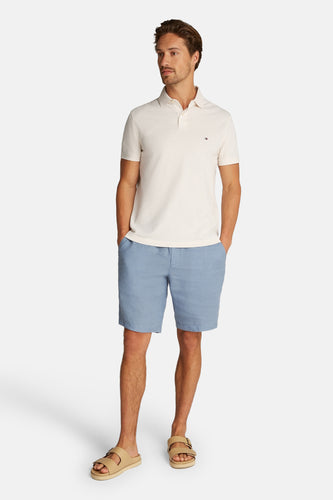 Man met beige polo, blauwe shorts en sandalen op witte achtergrond.

