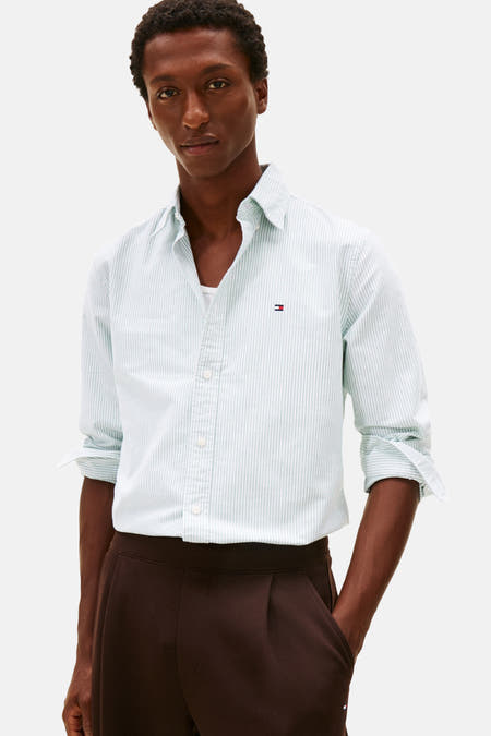 Chemise rayée verte de TOMMY HILFIGER à manches longues, portée avec un pantalon marron plissé et des manches retroussées.