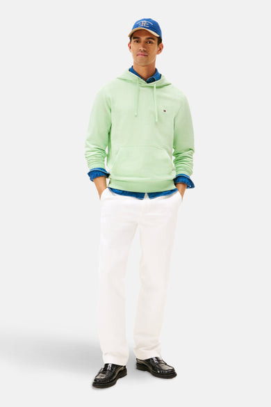 Pull-over vert de Tommy Hilfiger avec capuche, associé à une chemise bleue, un pantalon blanc et des chaussures noires, plus une casquette bleue.