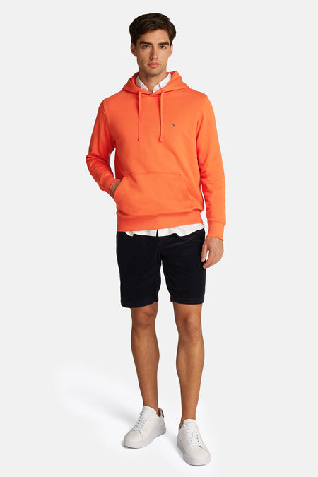 Pull-over orange Tommy Hilfiger avec capuche, combiné à une chemise blanche et un short noir.