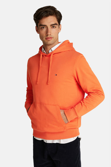 Pull-over orange Tommy Hilfiger avec capuche, cordons et poche, porté sur une chemise blanche.