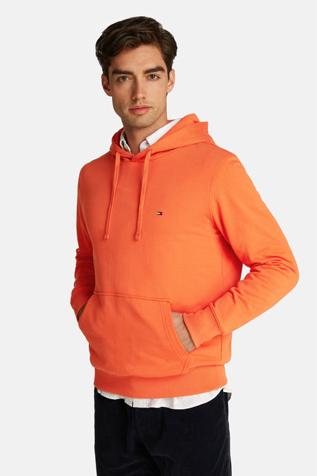 Pull-over orange Tommy Hilfiger avec capuche, cordons et poche, porté sur une chemise blanche.