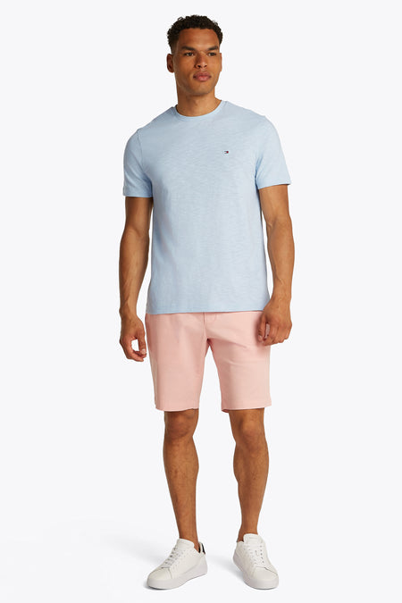 T-shirt à manches courtes bleu clair de TOMMY HILFIGER avec col rond et petit logo sur la poitrine, porté avec un short rose et des baskets blanches, vue de face.