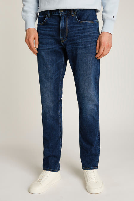 Jean droit modèle en denim bleu marine de TOMMY HILFIGER, porté avec des baskets blanches et une chemise bleue claire.