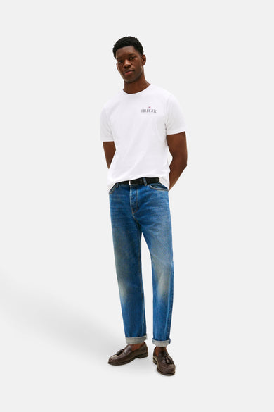 T-shirt blanc à manches courtes de TOMMY HILFIGER, porté avec des jeans bleus, une ceinture noire et des loafers marrons.