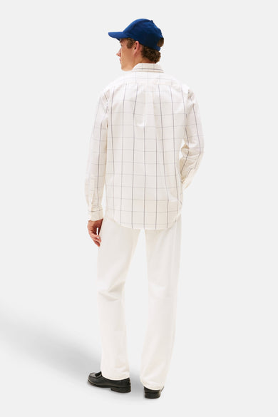 Man in wit hemd met ruitpatroon, witte broek en blauwe pet, staand van achteren op een witte achtergrond.