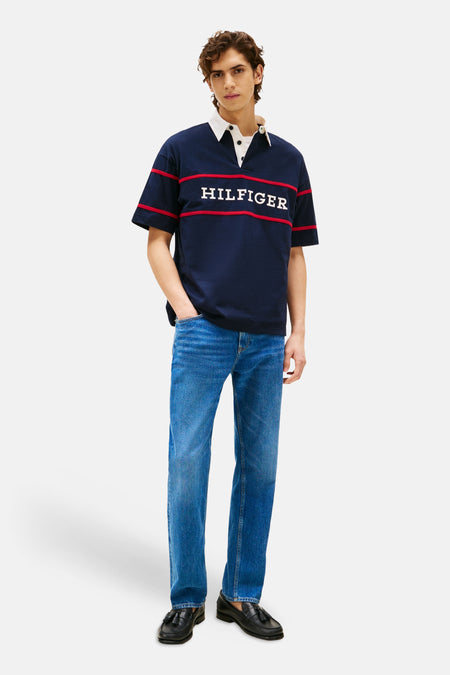 Polo bleu à manches courtes de Tommy Hilfiger, avec des rayures rouges et 'Hilfiger' en lettres blanches, porté avec un jean bleu et des mocassins noirs.