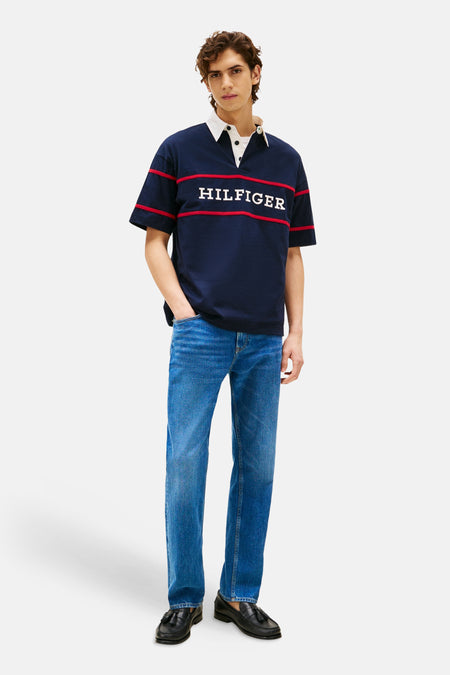 Polo bleu à manches courtes de Tommy Hilfiger, avec col blanc et rayures rouges et le texte 'HILFIGER'. Porté avec un jean bleu et des chaussures noires.
