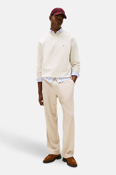 Sweater écru avec col rond de TOMMY HILFIGER, porté sur une chemise rayée, avec un pantalon beige et des chaussures marron.