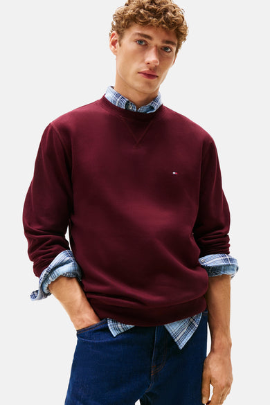 Pull-over rouge à col rond de TOMMY HILFIGER, porté sur une chemise à carreaux et des jeans bleus.