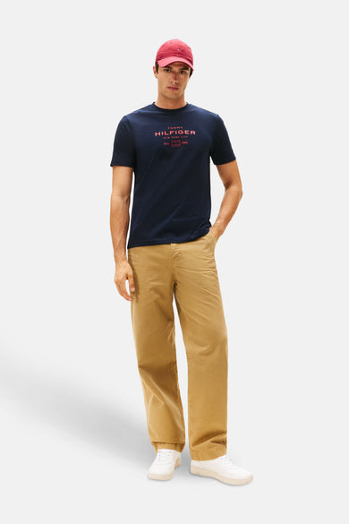T-shirt bleu à manches courtes de Tommy Hilfiger, avec une impression de texte rouge et porté avec un chapeau rouge et un pantalon kaki.