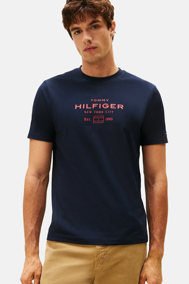 T-shirt bleu à manches courtes de TOMMY HILFIGER avec une impression rouge vive sur l'avant, porté avec un pantalon beige.