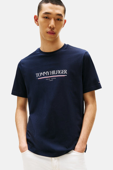 T-shirt bleu à manches courtes de TOMMY HILFIGER, vu de l'arrière, associé à un pantalon blanc et des chaussettes noires.