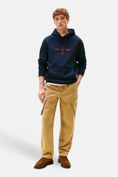 Pull-over bleu avec capuche de Tommy Hilfiger, porté avec un T-shirt blanc et un pantalon cargo kaki, avec des chaussures marrons.