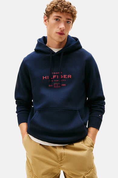 Pull-over bleu Tommy Tommyfiger avec un capuchon, avec un logo rouge sur la poitrine, porté avec un pantalon kaki.