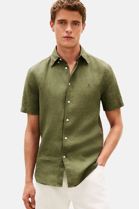Chemise verte de TOMMY HILFIGER, manches courtes, avec boutons blancs et logo brodé, combinée avec un pantalon blanc et des mocassins noirs.