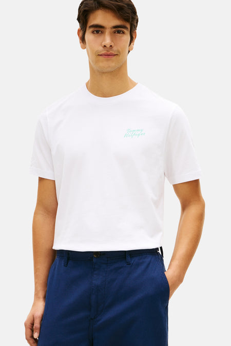 T-shirt blanc à manches courtes de TOMMY HILFIGER avec logo bleu clair sur la poitrine, porté par un homme avec un pantalon bleu foncé et des mocassins marron.