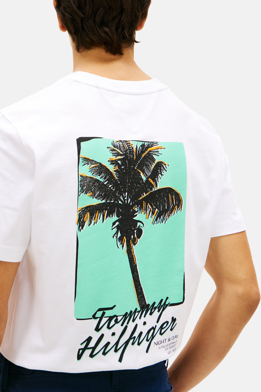 T-shirt blanc à manches courtes Tommy Hilfiger, avec un imprimé palmier et le logo de la marque dans le dos, porté par une personne.
