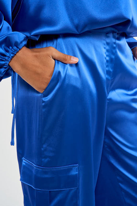 Pantalon cargo bleu de NOOR BY NORA GHARIB, avec une main dans la poche, en tissu brillant.