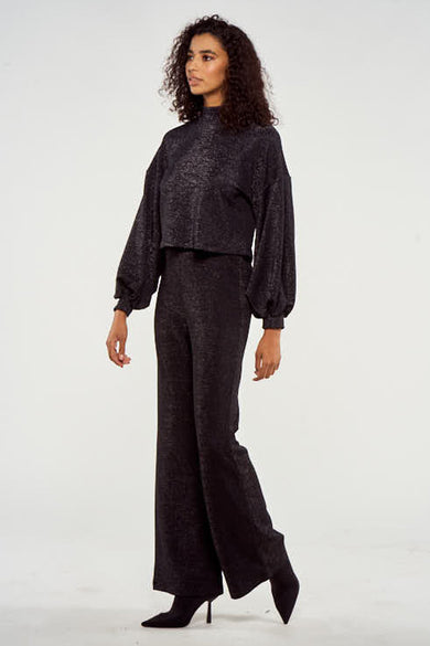 Pull noir à manches longues de NOOR BY NORA GHARIB, tissu brillant, porté avec un pantalon large et des bottes.