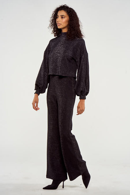 Pull noir à manches longues de NOOR BY NORA GHARIB, tissu brillant, porté avec un pantalon large et des bottes.