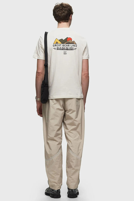 T-shirt écru de Napapijri, avec des montagnes et un logo sur le dos, associé à une pantalon beige et une sac noir.
