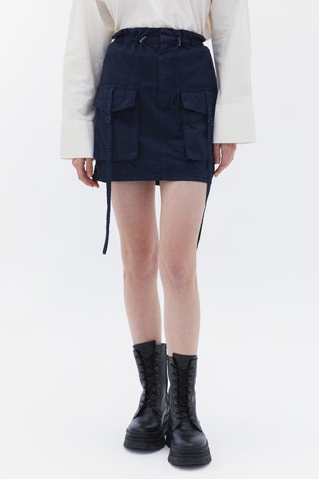 Jupe courte bleue OXXO, avec poches et cordon, associée à une blouse blanche et des bottes noires.