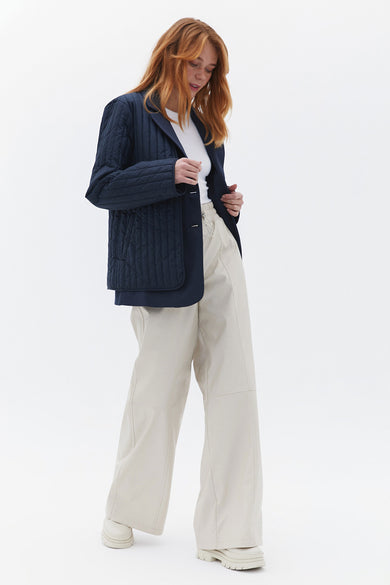 Manteau court bleu d'OXXO, avec un motif brodé, porté sur une chemise blanche et un pantalon beige.
