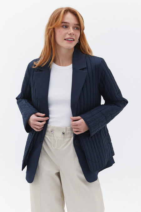 Manteau court bleu d'OXXO, avec un motif de lunettes à carreaux et porté sur une chemise blanche et un pantalon beige.