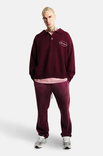 Pull à col V rouge de ANOTHER COTTON LAB, avec une badge brodée, associé à un pantalon de jogging bordeaux et des baskets grises.