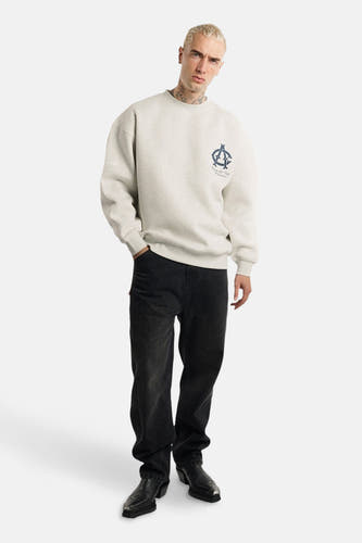 Pull-over gris d'ANOTHER COTTON LAB, avec un petit logo bleu marine sur la poitrine, porté avec des jeans noirs et des bottes de cow-boy.