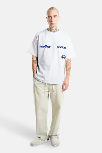 T-shirt blanc à manches courtes d'ANOTHER COTTON LAB, avec une impression de texte bleue et porté avec un pantalon décontracté clair.